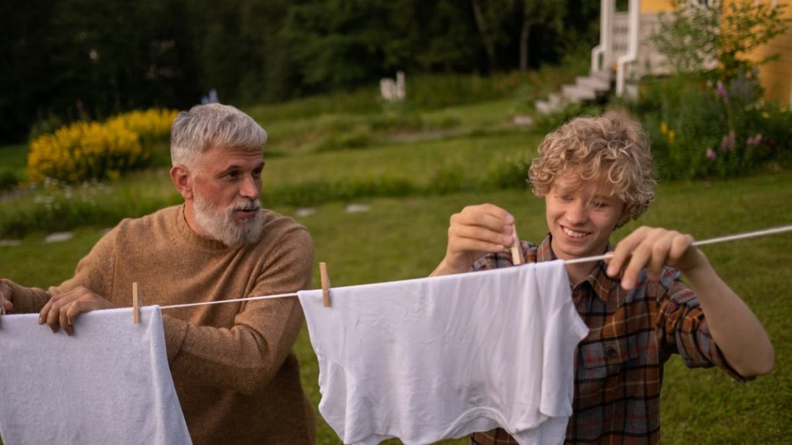 Elderly man and teenage boy joyfully hanging laundry in a sunny garden setting.